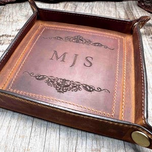 May include: A square, brown leather valet tray with a stitched border and brass button details. The centre features the engraved initials "MJS" above and below decorative scrollwork.