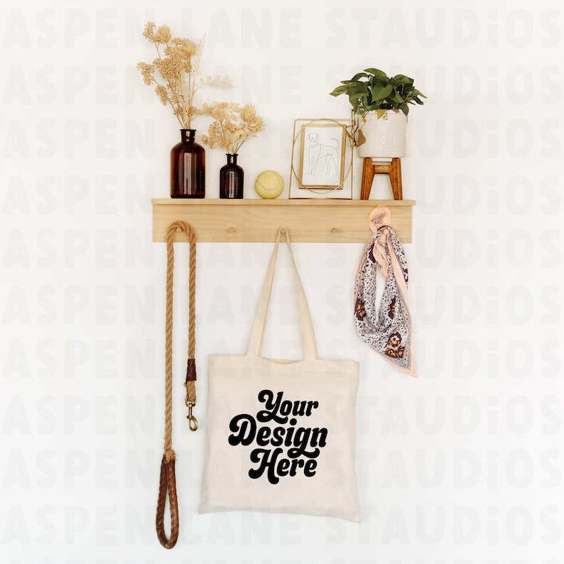 Puede incluir: Una bolsa de tela blanca con texto negro que dice "Your Design Here" colgada de una estanter&iacute;a de madera con una correa de cuerda, un jarr&oacute;n de vidrio marr&oacute;n con flores secas, una peque&ntilde;a botella de vidrio, una pelota amarilla, un marco de fotos dorado, una planta en maceta y una bufanda rosa.
