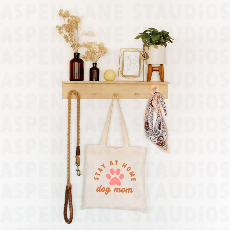 Puede incluir: Una bolsa de lona blanca con el texto "Stay at Home dog mom" en naranja y una huella de pata rosa. La bolsa est&aacute; colgada en un estante de madera con una correa de cuerda marr&oacute;n, una peque&ntilde;a planta en maceta y una imagen enmarcada.