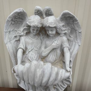 May include: Two white painted angel figurines with wings, sitting inside a large white seashell. The angels are holding hands and looking down.