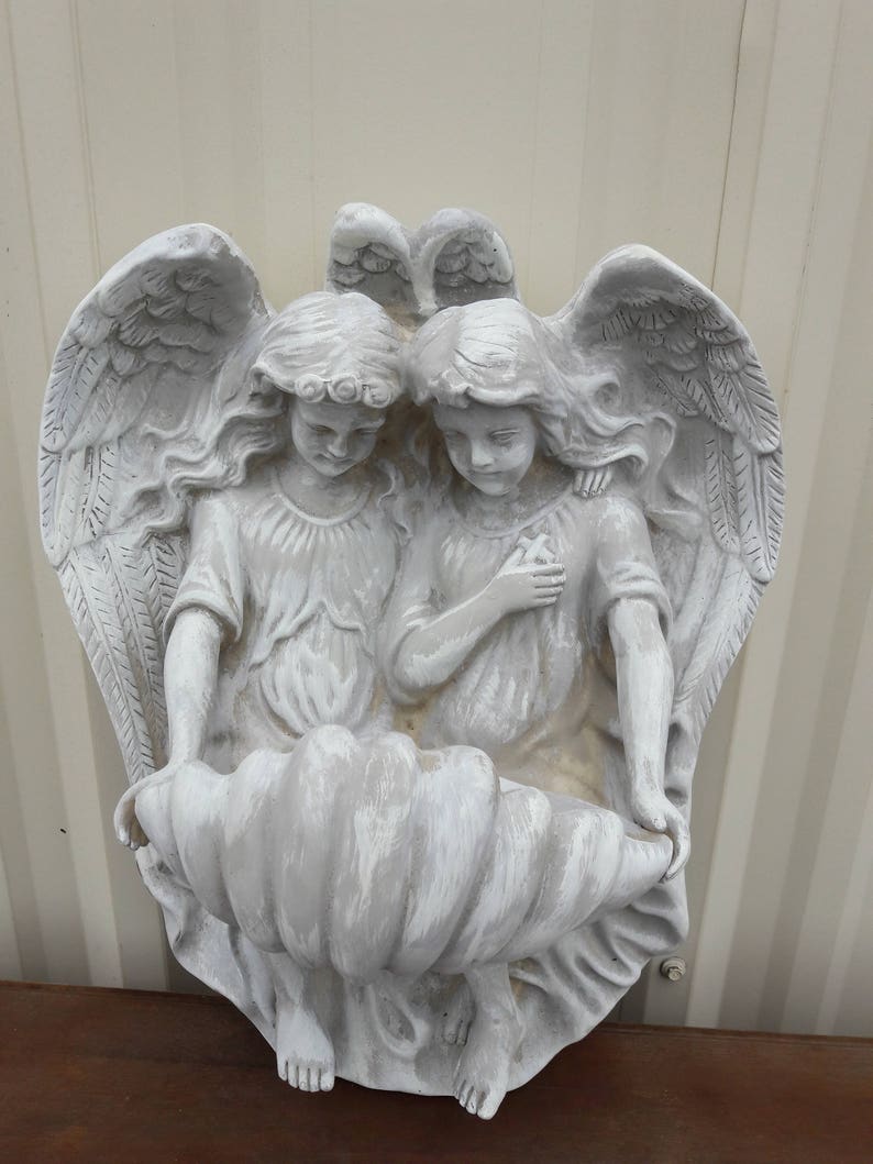 May include: Two white painted angel figurines with wings, sitting inside a large white seashell. The angels are holding hands and looking down.