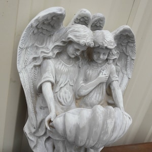 May include: Two white painted angel figurines with wings, holding a large white seashell planter. The angels are facing each other and have long flowing hair.