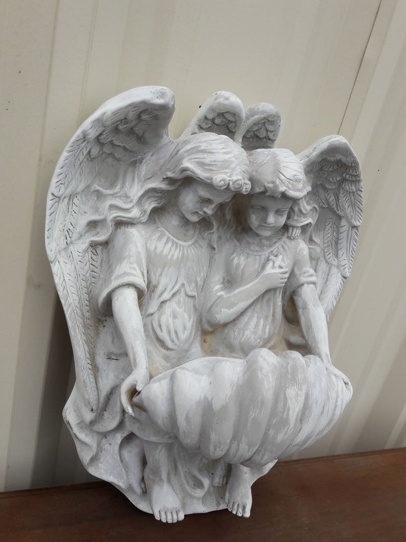 May include: Two white painted angel figurines with wings, holding a large white seashell planter. The angels are facing each other and have long flowing hair.