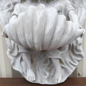 May include: White painted angel statue holding a large, white, shell-shaped planter. The angel has wings and is holding the planter in its hands.