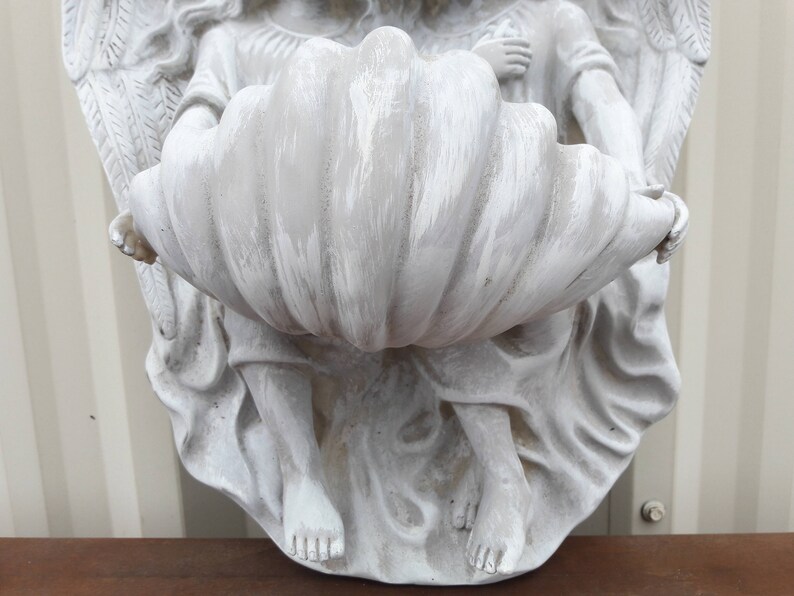 May include: White painted angel statue holding a large, white, shell-shaped planter. The angel has wings and is holding the planter in its hands.