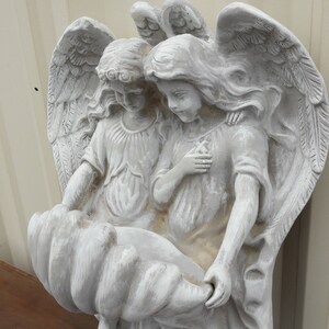 May include: A white painted plaster sculpture of two angels holding a bird's nest. The angels are depicted with wings and long flowing hair. The sculpture is a wall hanging.