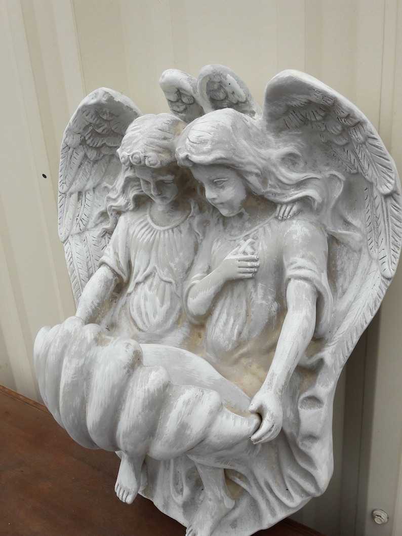 May include: A white painted plaster sculpture of two angels holding a bird's nest. The angels are depicted with wings and long flowing hair. The sculpture is a wall hanging.