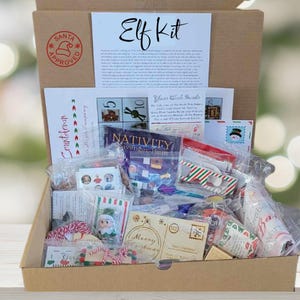 May include: A cardboard box containing an Elf Kit with various holiday-themed items in plastic bags. The box lid features a Santa Approved stamp and a printed letter titled Elf Kit. Inside are activity cards, a Nativity scavenger hunt, and small toys.