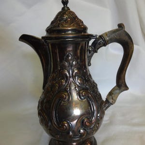 May include: A silver coffee pot with an ornate design and a wooden handle. The pot has a round body with a spout and a lid. The handle is attached to the side of the pot and is made of wood.