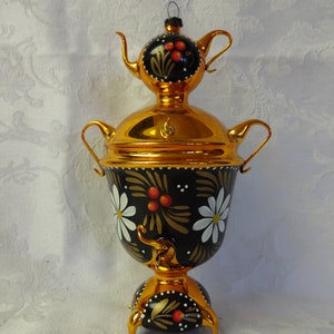 May include: A traditional Russian samovar with a black and gold painted design featuring white daisies and red berries. The samovar has a gold-colored base with three legs and a gold-colored lid.