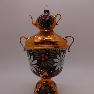 May include: A decorative, gold-colored samovar ornament with a black base. The ornament features white daisy-like flowers and red berries. The top has a small spout and handle. The samovar sits on three gold legs.