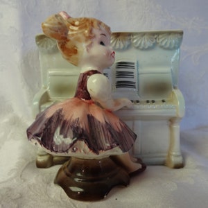 May include: A ceramic figurine of a girl playing a white piano. The girl wears a pink and purple dress and has blonde hair. The piano has black keys and a music sheet. The figurine is on a brown base.