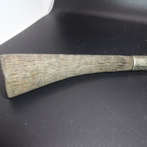 May include: Close-up of a vintage knife handle. The handle is made of textured, light brown material with a silver band and decorative etching near the blade. The blade is a dark metal.