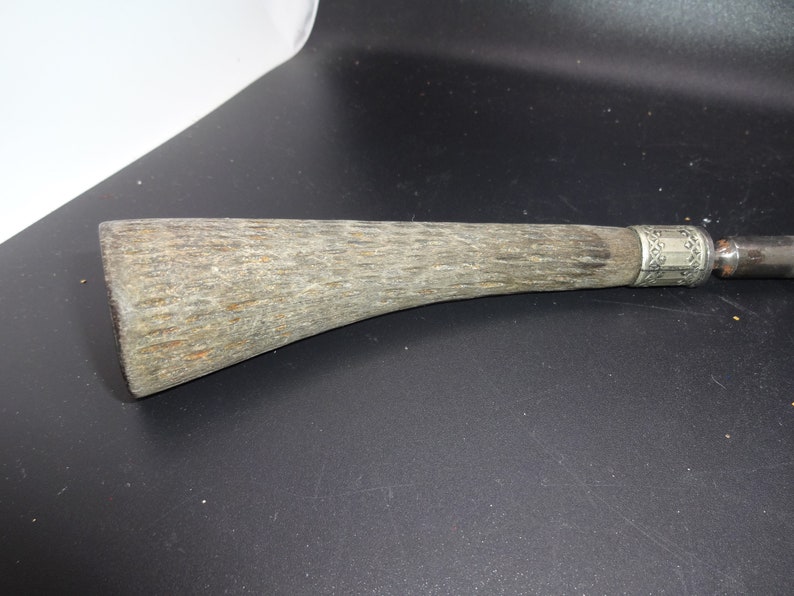 May include: Close-up of a vintage knife handle. The handle is made of textured, light brown material with a silver band and decorative etching near the blade. The blade is a dark metal.