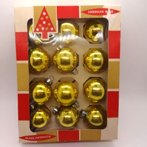 May include: A vintage box of twelve gold-colored Christmas ornaments. The ornaments are round and shiny, reflecting the surrounding environment. The box is beige with red and gold accents, and the text "AMERICAN MADE" and "GLASS PRODUCTS" are visible.