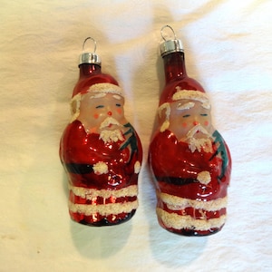 May include: Two vintage glass Christmas ornaments shaped like Santa Claus. The ornaments are red with white trim and have a green and white painted design.