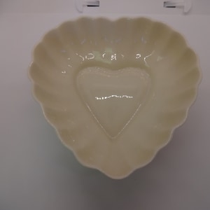May include: Cream-colored heart-shaped dish with a scalloped edge. The center of the dish features a heart-shaped indentation. The dish is made of a glossy material and is suitable for serving or display.