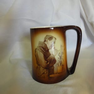 May include: A brown ceramic mug with a handle. The mug features a painting of a man in a brown robe holding a plant. The plant is in a brown pot. The mug has a brown glaze.