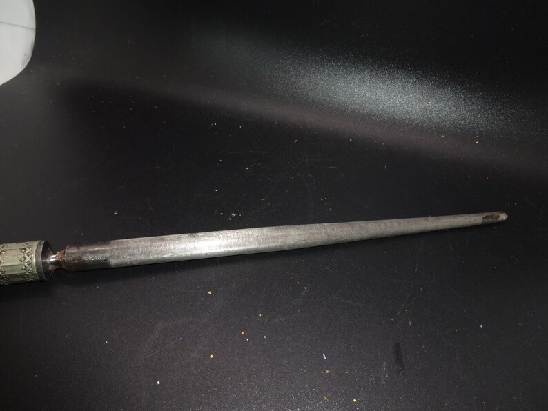 May include: A silver-toned metal object with a long, tapered point. The handle has a decorative, patterned design. The object appears to be a tool, possibly for sharpening or piercing.