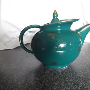 May include: A teal teapot with a gold lid and gold floral accents. The teapot has a round body and a curved spout.