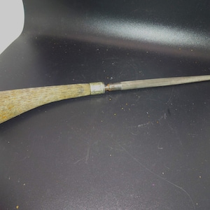May include: A steel knife sharpener with a light-colored, textured handle. The handle is curved and attached to a long, tapered steel rod. A silver band connects the handle to the steel rod. The sharpener is designed for sharpening knives.