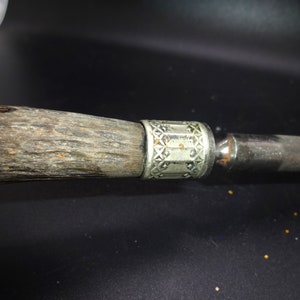 May include: Close-up of a knife handle and blade. The handle is made of textured, dark wood with a silver-colored metal band featuring a geometric pattern. The blade is a polished, silver color.