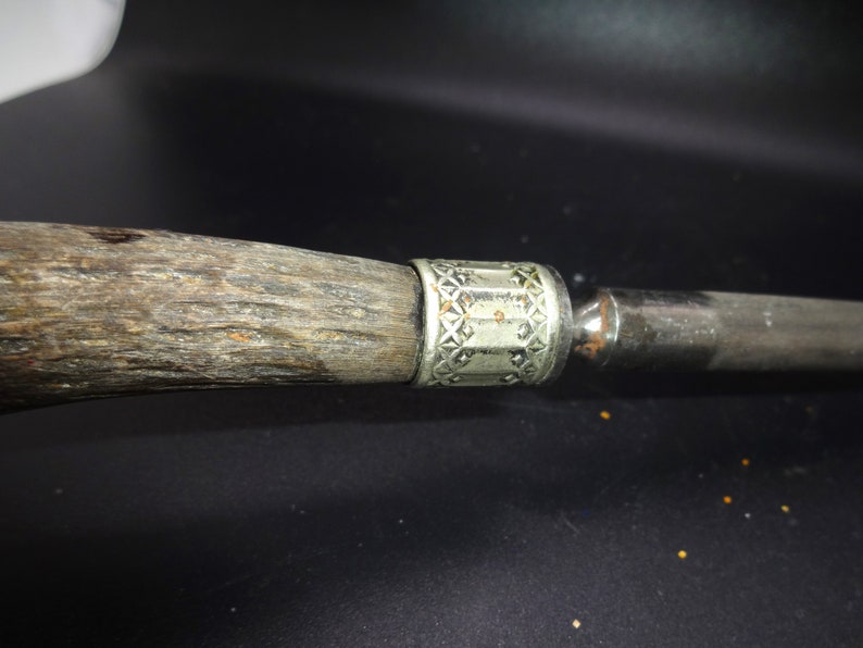 May include: Close-up of a knife handle and blade. The handle is made of textured, dark wood with a silver-colored metal band featuring a geometric pattern. The blade is a polished, silver color.