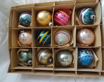 May include: A vintage box of Christmas ornaments. The ornaments are round and come in various colors, including gold, blue, pink, and silver. Some have stripes, glitter, or other decorative patterns. Each ornament is nestled in its own compartment.