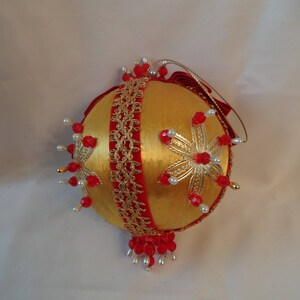 May include: A gold Christmas ornament with red and white beads and a gold ribbon. The ornament is decorated with a gold and white snowflake design.