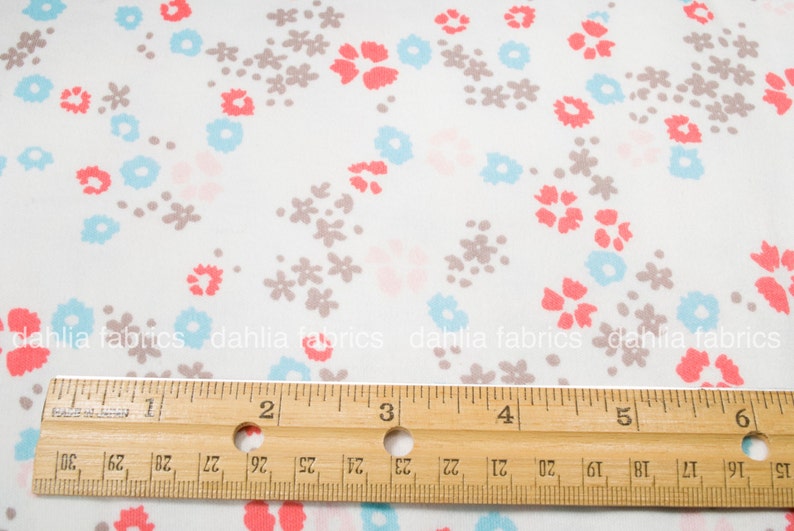 May include: White fabric with a repeating pattern of small pink, blue, and gray flowers. A wooden ruler is placed on top of the fabric, with the numbers 1 through 6 visible.