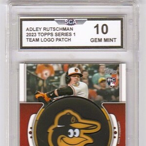 May include: A baseball card featuring Adley Rutschman, a player for the Baltimore Orioles. The card is graded Gem Mint 10 by the Sportscard Guaranty Corporation (SCG). The card features a team logo patch and a photo of Rutschman in his Orioles uniform.