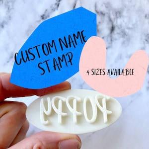 Puede incluir: Un sello de nombre personalizado blanco de forma ovalada con el nombre "OFF" en una fuente estilizada. El sello está siendo sostenido por una mano. Dos notas de papel de colores están en el fondo, una azul con el texto "CUSTOM NAME STAMP" y una rosa con el texto "4 SIZES AVAILABLE".