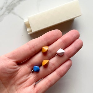May include: Four small, colourful, diamond-shaped resin pieces in a hand. The pieces are blue, orange, orange, and white.