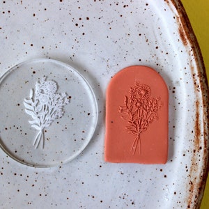 May include: A clear polymer stamp with a floral design, and a red polymer stamp with the same floral design. The stamps are on a white speckled surface.