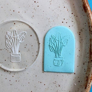 May include: A clear acrylic stamp and a blue silicone stamp, both featuring a line drawing of a potted plant with leaves.