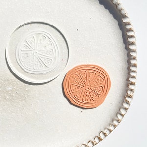 May include: A white ceramic plate with a raised edge and a decorative pattern. On the plate are two orange-coloured clay stamps, each with a raised design of a lemon slice.