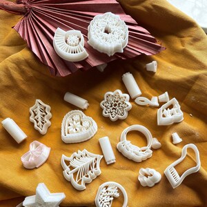 Puede incluir: Una colección de cortadores de galletas de plástico blanco en varias formas, incluyendo una estrella, un árbol, un corazón, un copo de nieve, una corona, una campana y una media. Los cortadores están dispuestos sobre un fondo de tela amarilla.