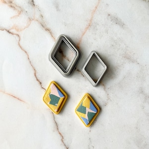 May include: Two pairs of colorful, geometric earrings with a yellow, green, blue, and pink design. The earrings are made of polymer clay and have a gold rim. The image also shows two sets of square-shaped cookie cutters.