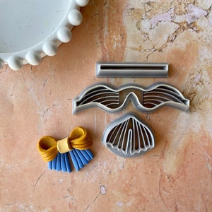 Puede incluir: Un juego de cuatro cortadores de galletas de plata, que incluyen un lazo, unas gafas de sol, una concha y un rectángulo. Los cortadores de galletas están sobre una superficie marrón claro.