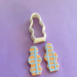 Pode incluir: Dois brincos de argila polimérica com padrão azul claro e laranja em forma de figura ondulada e arredondada. Também é mostrado um cortador de biscoitos de plástico branco com a forma dos brincos.