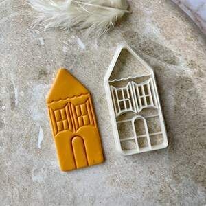 Può includere: Un set di due stampini per biscotti a forma di case con finestre e porte. Gli stampini sono in plastica bianca e sono mostrati su una superficie marrone chiaro.