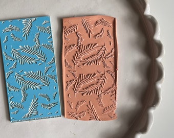 以下が含まれることがあります： 葉の模様がついた長方形のシリコンスタンプが2つ。1つは青、もう1つはオレンジです。