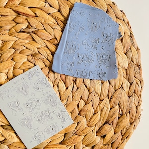 May include: Two gray rubber stamps with a repeating pattern of a tiger's face. The stamps are on a woven straw surface.
