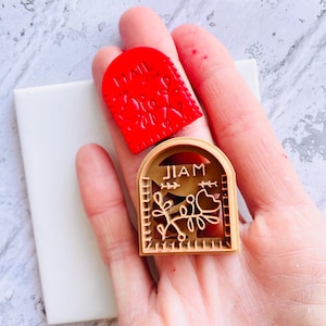 May include: A red, arched-top stamp with a floral design and the word "MAIL" is next to a wooden stamp with the word "JIAM" and a floral and dog design. The stamps are on a white surface.
