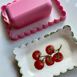 May include: A rectangular ceramic dish with a scalloped edge, featuring a hand-painted vine of red cherry tomatoes on a white speckled background, paired with a matching pink plastic rectangular scalloped cookie cutter.