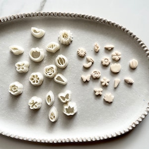 May include: A white ceramic plate with two sets of small, white, plastic cookie cutters in the shapes of flowers, leaves, butterflies, and other designs.