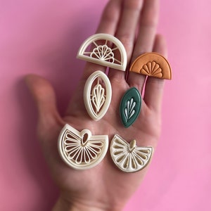 May include: A set of seven white, green and orange polymer clay cookie cutters shaped like half circles, leaves and lemons. The cutters are arranged on a hand.