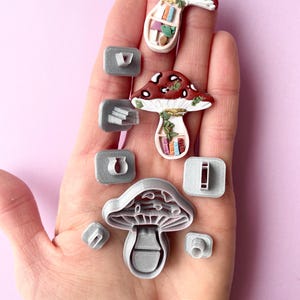 Può includere: Un set di otto stampini per biscotti in plastica grigia a forma di fungo. Due degli stampini sono più grandi e presentano un design di fungo dettagliato con un gambo e un cappello. Gli altri sei stampini sono più piccoli e presentano varie forme, tra cui un rettangolo, un ferro di cavallo e un triangolo.