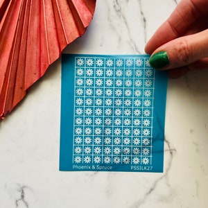 Puede incluir: Una hoja azul con una cuadrícula de margaritas blancas. La hoja tiene el texto "Phoenix & Spruce" y "PSSILK27" impreso.