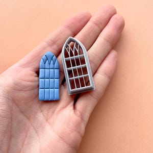 May include: Two gray plastic molds for making fondant or gum paste window shapes. The mold on the left is a solid blue window shape. The mold on the right is a detailed window frame.